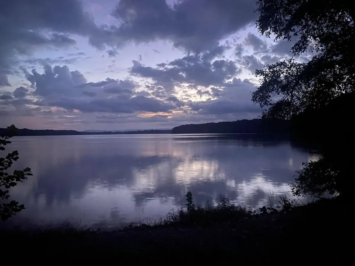 A tranquil view of Tims Ford Lake, showcasing calm waters, a cloudy sky, and a distant tree line. The scene captures the essence of lakeside activities such as boating, kayaking, and fishing, while also inviting peaceful reflection by the water.