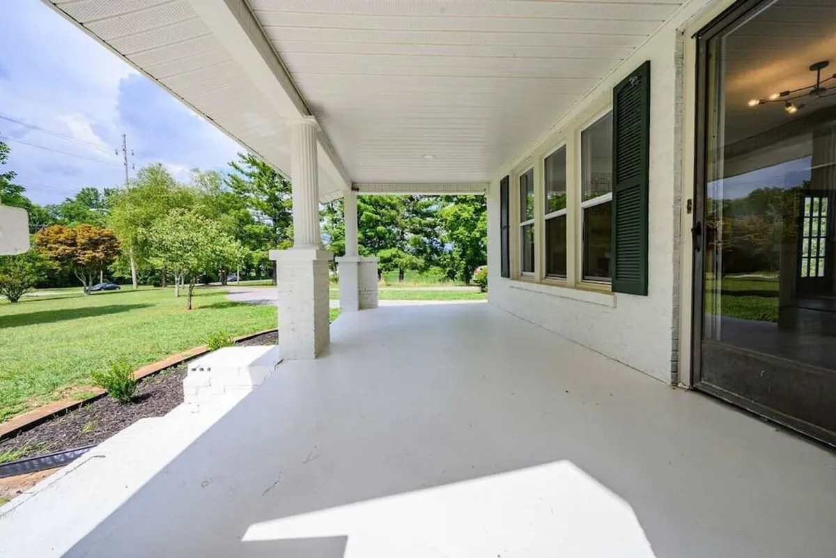 A wide view of the home's large porch, set amidst lush green landscaping and featuring a glass-paneled front door. The porch is staged for relaxation—ideal for enjoying morning coffee, waving to neighbors, or hosting intimate gatherings.