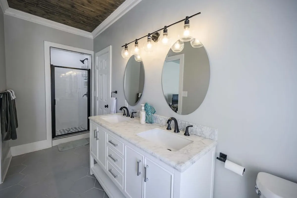 A spacious, modern bathroom with a double vanity featuring a marble countertop, large mirrors, stylish light fixtures, and a glass shower enclosure. The space is bright, clean, and evokes a spa-like feel.