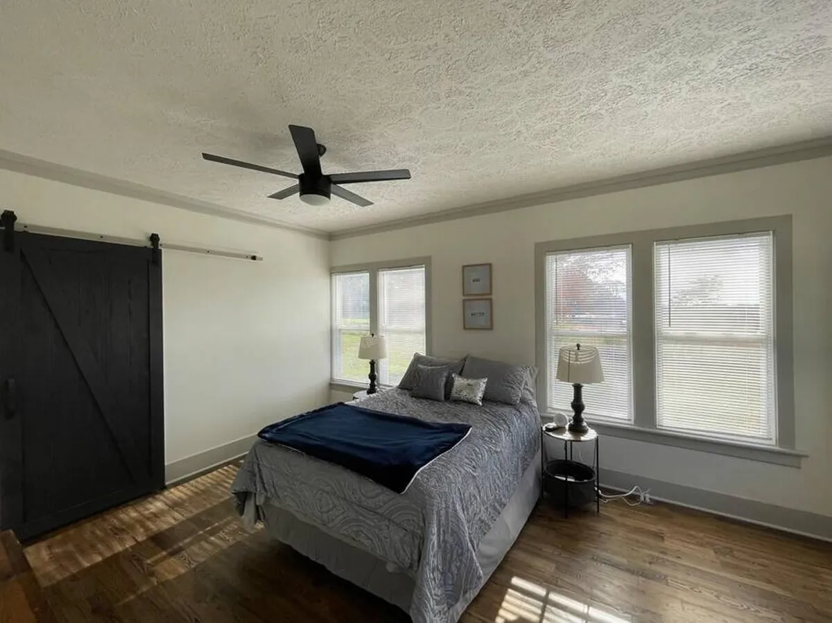 A bedroom scene featuring a bed, windows, a barn door, and a nightstand with a lamp. Gentle natural light fills the room, creating a calm and serene setting suitable for reading or resting.