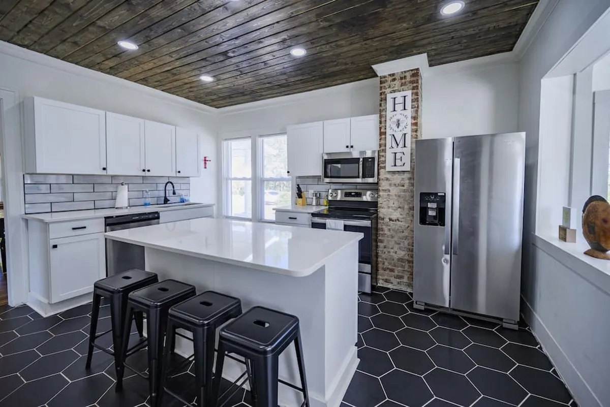 A stylish, functional kitchen with an island countertop, black stools for casual seating, exposed brick wall with a 'HOME' sign, and stainless steel appliances. The space is clearly designed for cooking, entertaining, and spending time together.