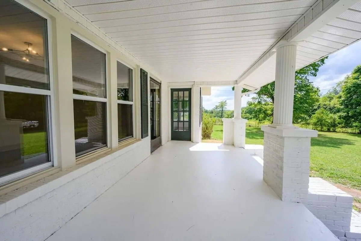 A spacious porch with classic columns overlooks a lush expanse of greenery, bathed in soft morning light. The setting suggests a tranquil spot to watch the sunrise, with surrounding fields and open air creating a serene atmosphere.